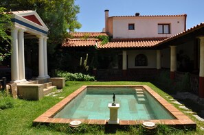Una piscina amb una estàtua al centre, envoltada d'un jardí amb escales que condueixen a una zona coberta.