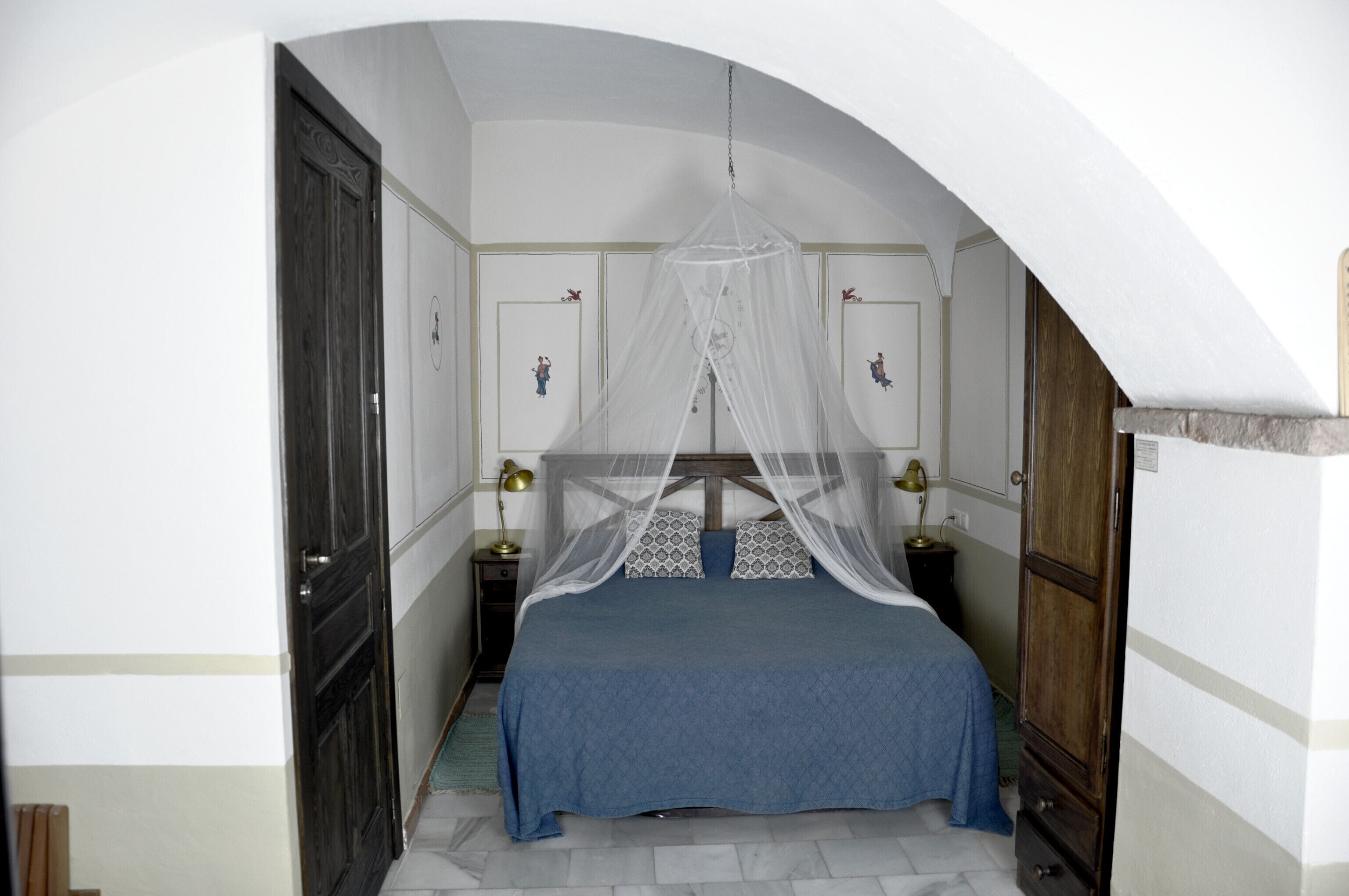 Een slaapkamer met een bed onder een baldakijn, twee nachtkastjes met lampen, en een deur aan de linkerkant.