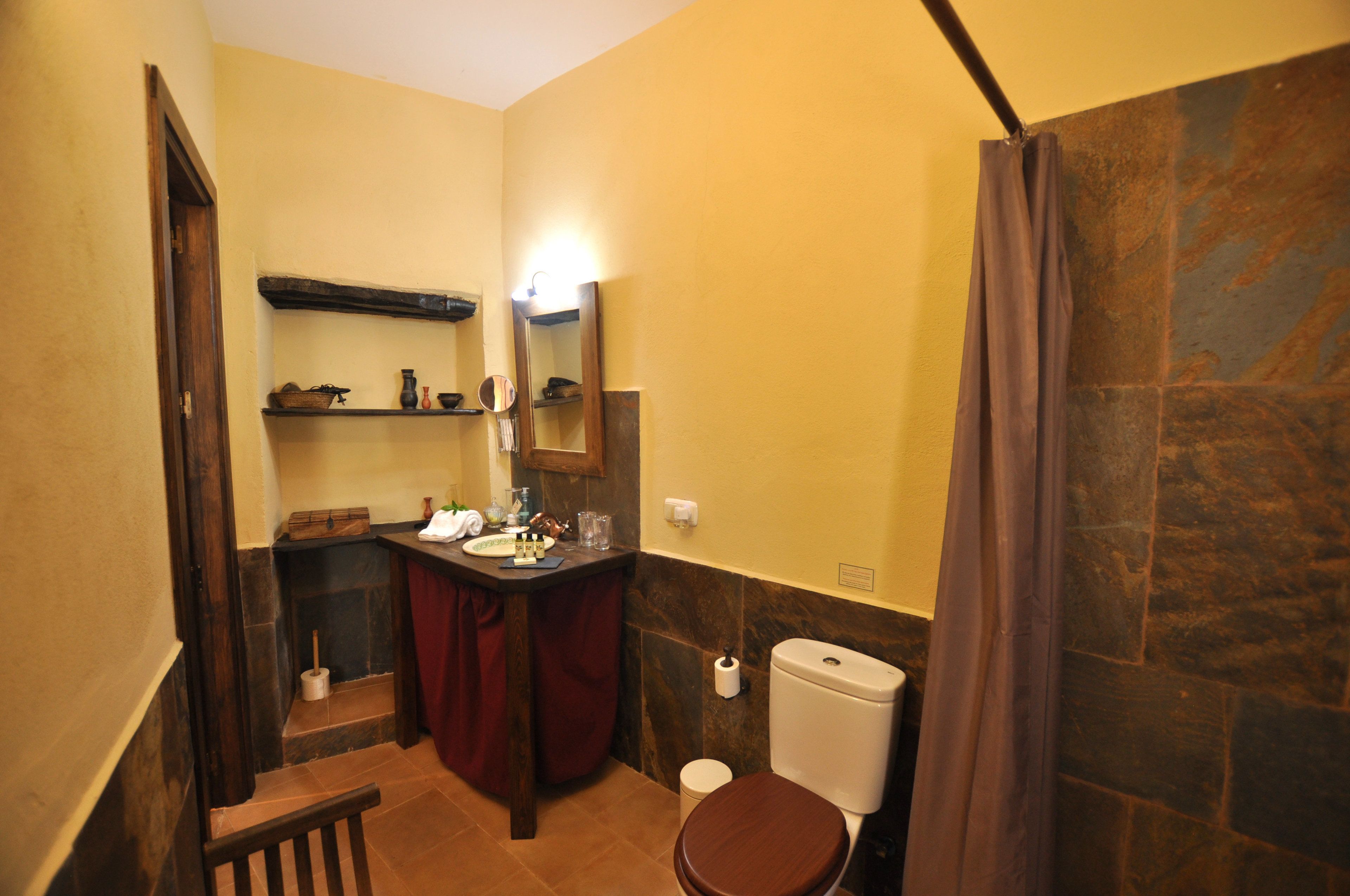 A bathroom with a wooden bench, a toilet, a sink with a mirror, and a shelf with toiletries.