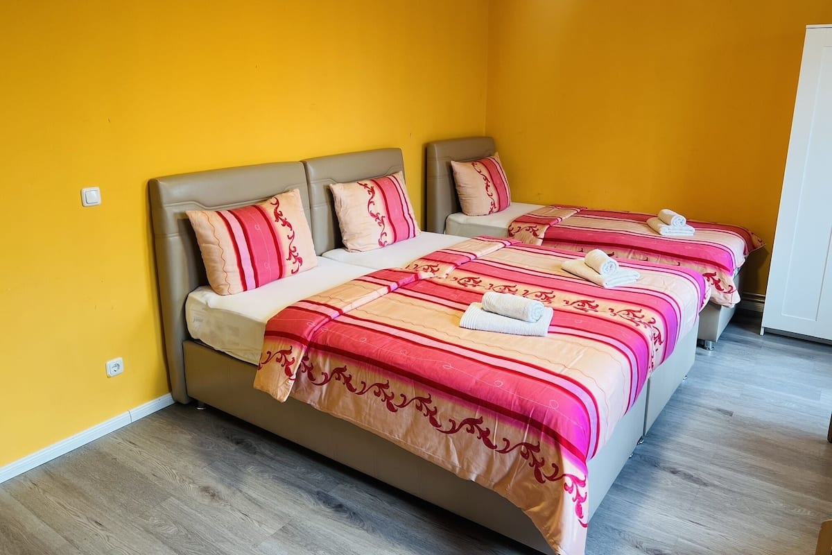 Een hotelkamer met twee bedden, elk voorzien van identiek rood en beige beddengoed, en een eenpersoonsbed met vergelijkbaar beddengoed.