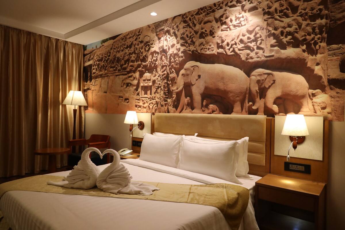 A hotel room with a large bed, two wall-mounted lamps, a nightstand, a chair, and a mural of elephants on the wall.