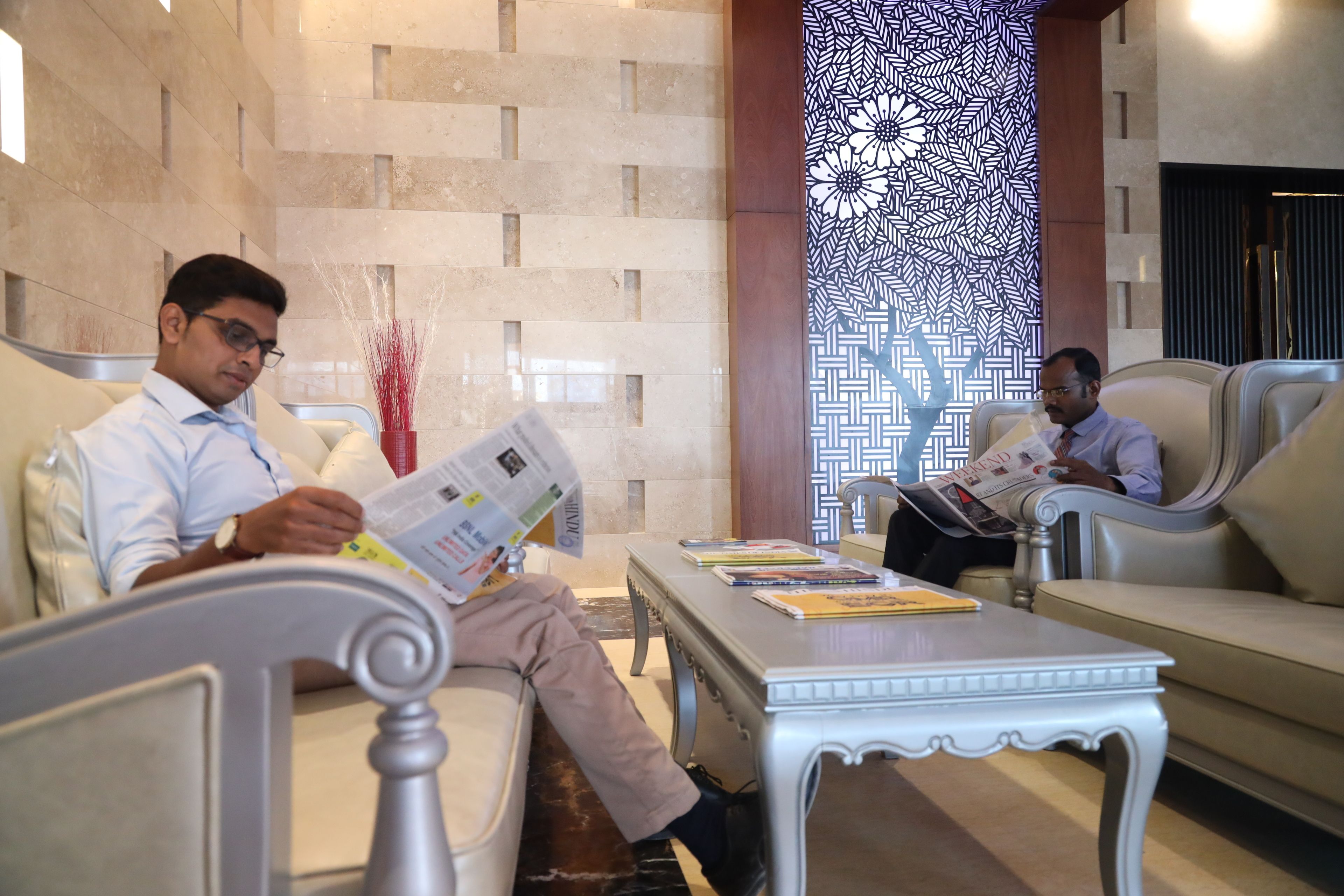 A modern hotel lobby with two men seated on sofas, reading newspapers. The lobby features a patterned wall and a decorative column.