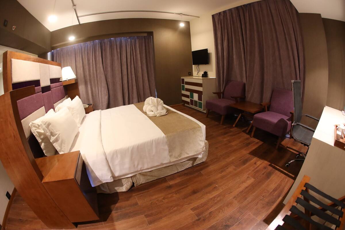 A hotel room with a large bed, a desk, a chair, a TV, and a desk lamp.