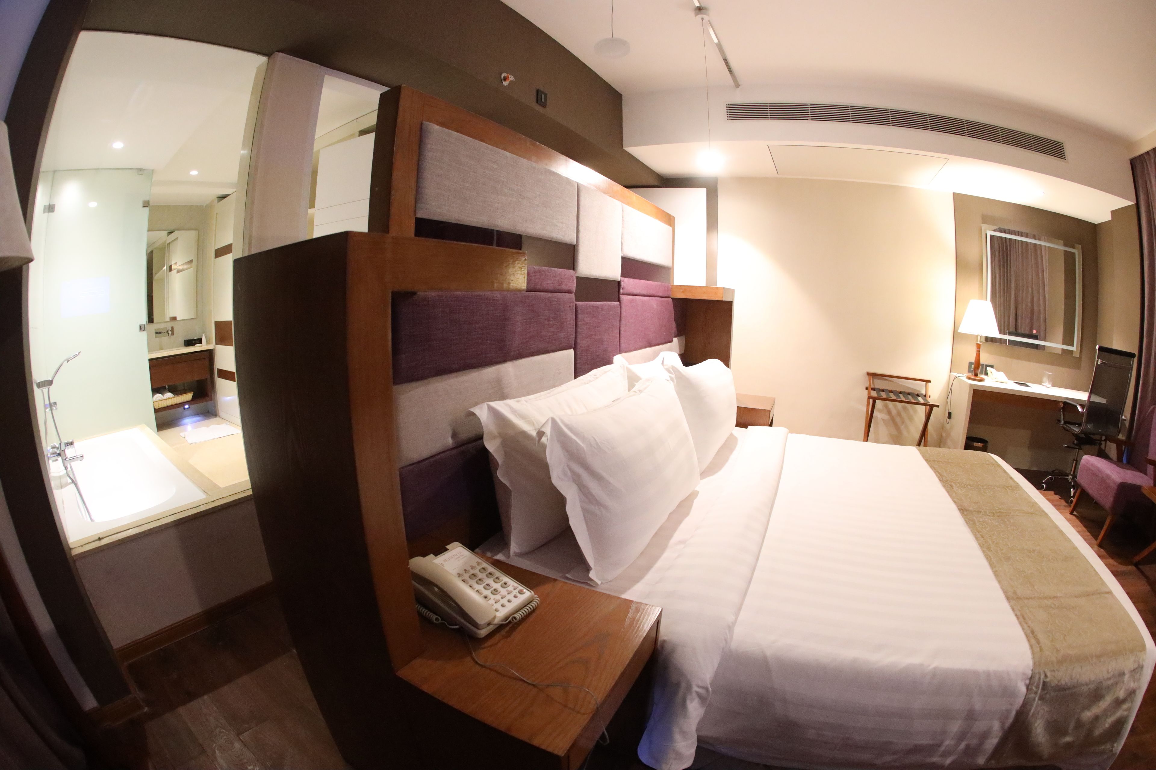 Una habitación de hotel con una cama grande, una mesita de noche de madera con un teléfono, un espejo y un escritorio con una lámpara.