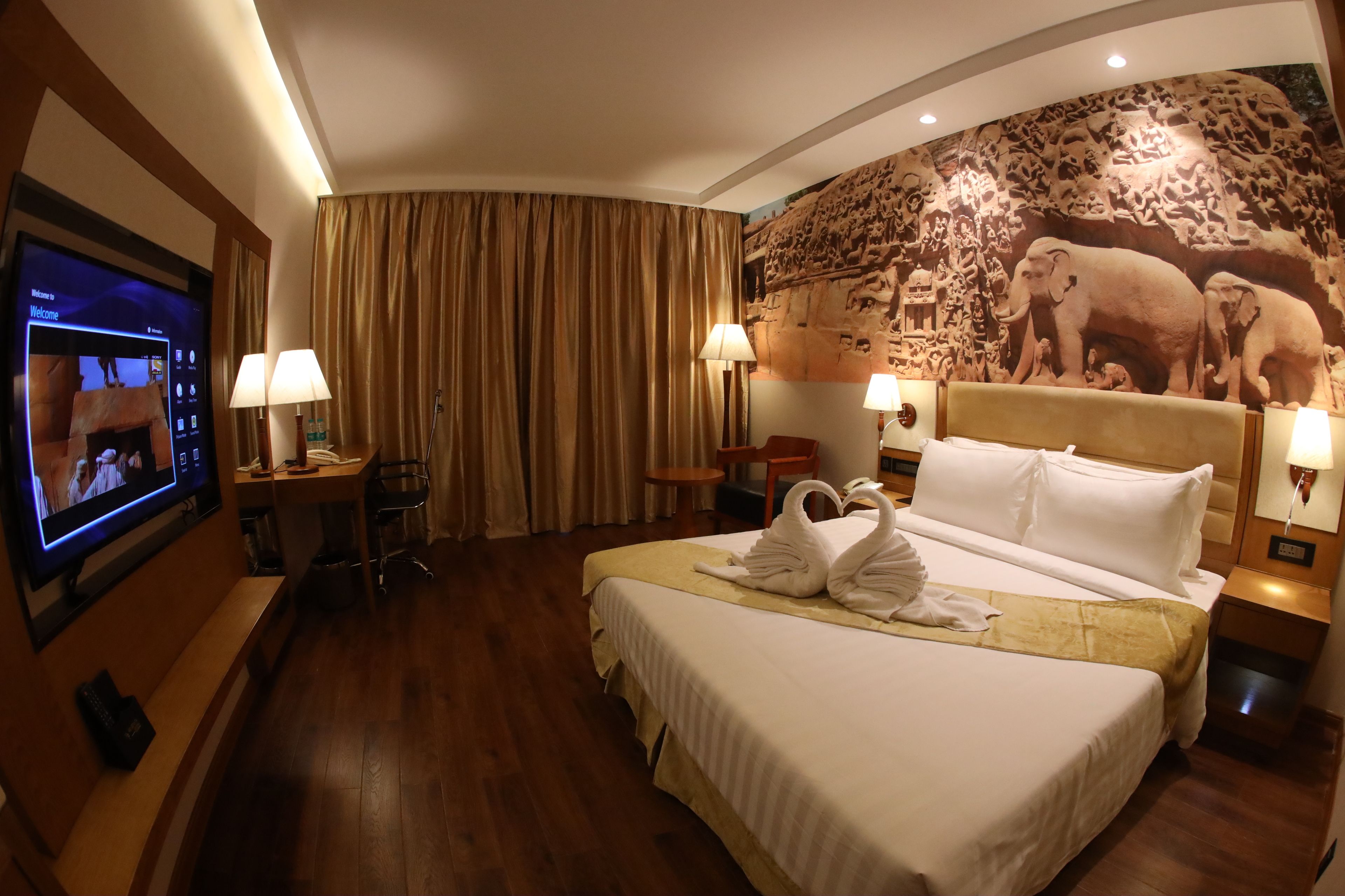 A hotel room with a large bed, a flat-screen TV, a desk with a lamp, and a mural of elephants on the wall.