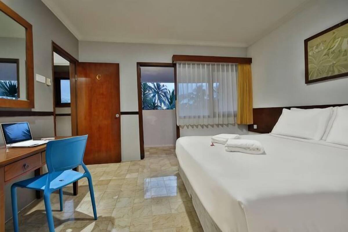 A hotel room with a bed, a desk with a laptop, a chair, a window with curtains, and a door leading outside.