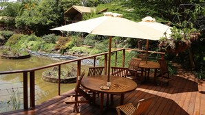 Una terraza de madera con una mesa redonda y sillas, un gran paraguas y vistas a un estanque con plantas y una cascada pequeña.