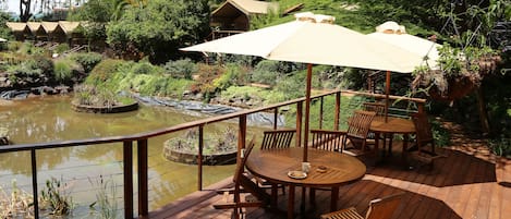 Una terraza de madera con una mesa redonda y sillas, un gran paraguas y vistas a un estanque con plantas y una cascada pequeña.