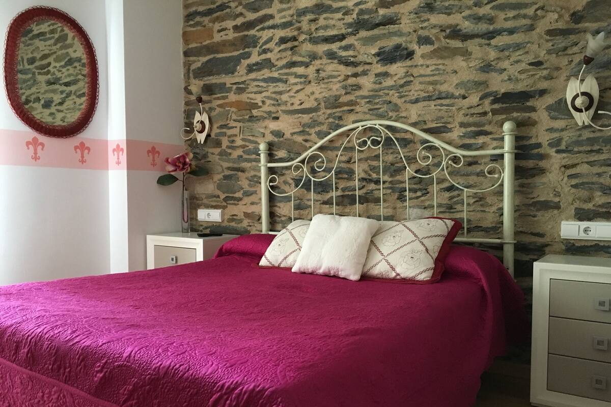 A bedroom with a stone wall, a metal bed frame, a pink bedspread, and a white nightstand with a lamp.