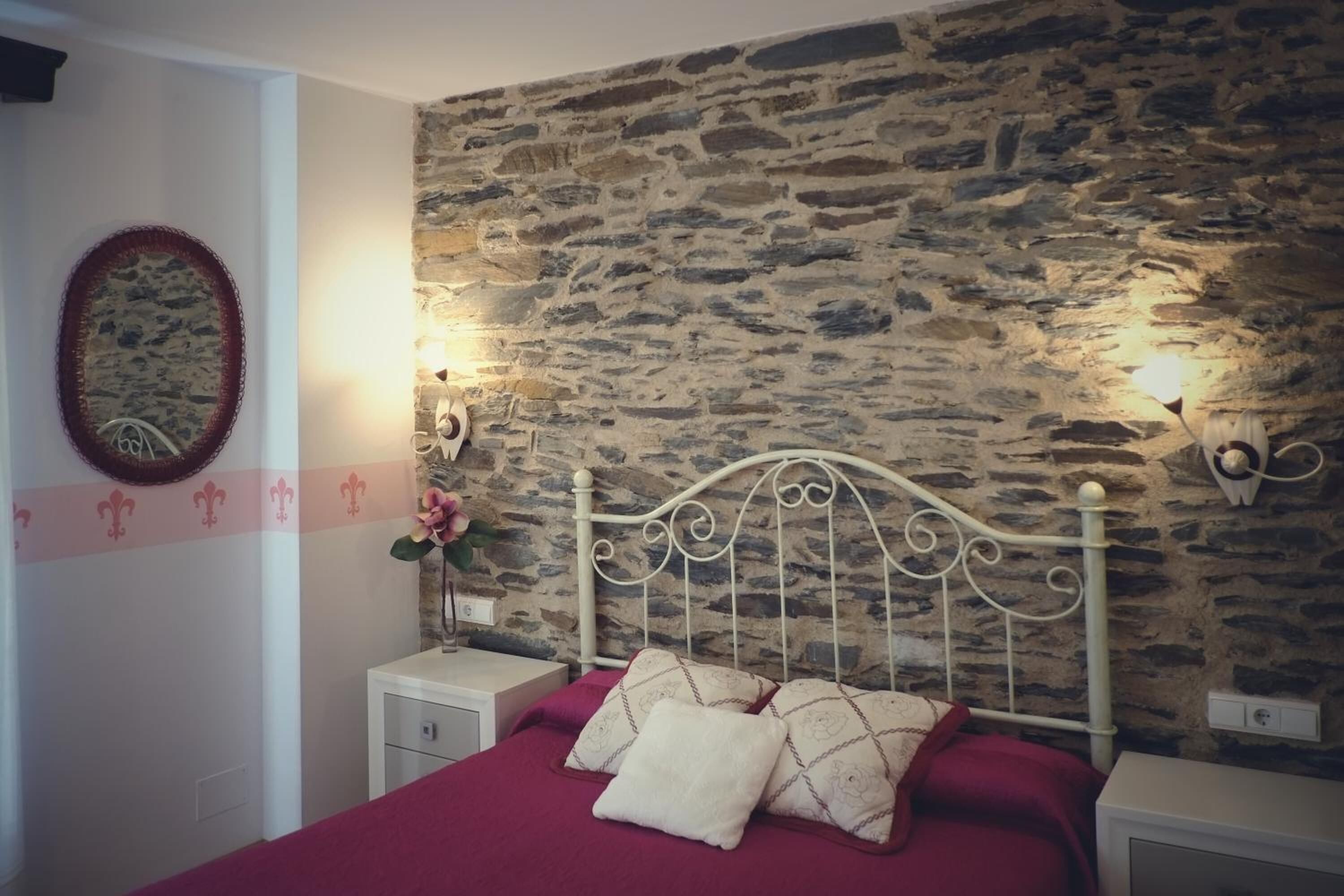A bedroom with a stone wall, a white metal bed frame, a red bedspread, and a nightstand with a lamp.