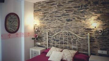 A bedroom with a stone wall, a white metal bed frame, a red bedspread, and a nightstand with a lamp.