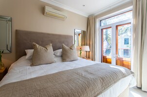 A neatly made bed with a beige headboard and pillows, a bedside table with a lamp, and a window with curtains.