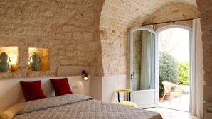 A bedroom with a stone wall, a bed with a quilted cover, two red pillows, a small table with a green vase, and a chair outside. - Masseria Grofoleo (Locorotondo)