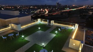 Una terraza en la azotea con césped verde, sillas y mesas blancas, todo rodeado por una barandilla blanca.
