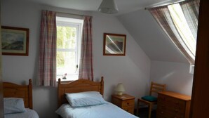 A bedroom with two single beds, a wooden dresser, a chair, a nightstand with a lamp, and a window with curtains. - Kirkwood Holiday Cottages (Lockerbie)