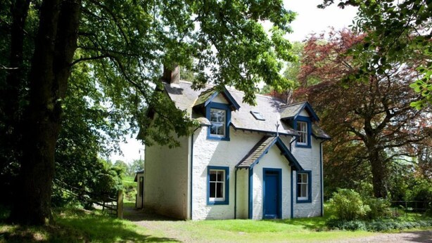 A quaint, single-story house with a gabled roof, surrounded by lush greenery and trees. - Kirkwood Holiday Cottages (Lockerbie)
