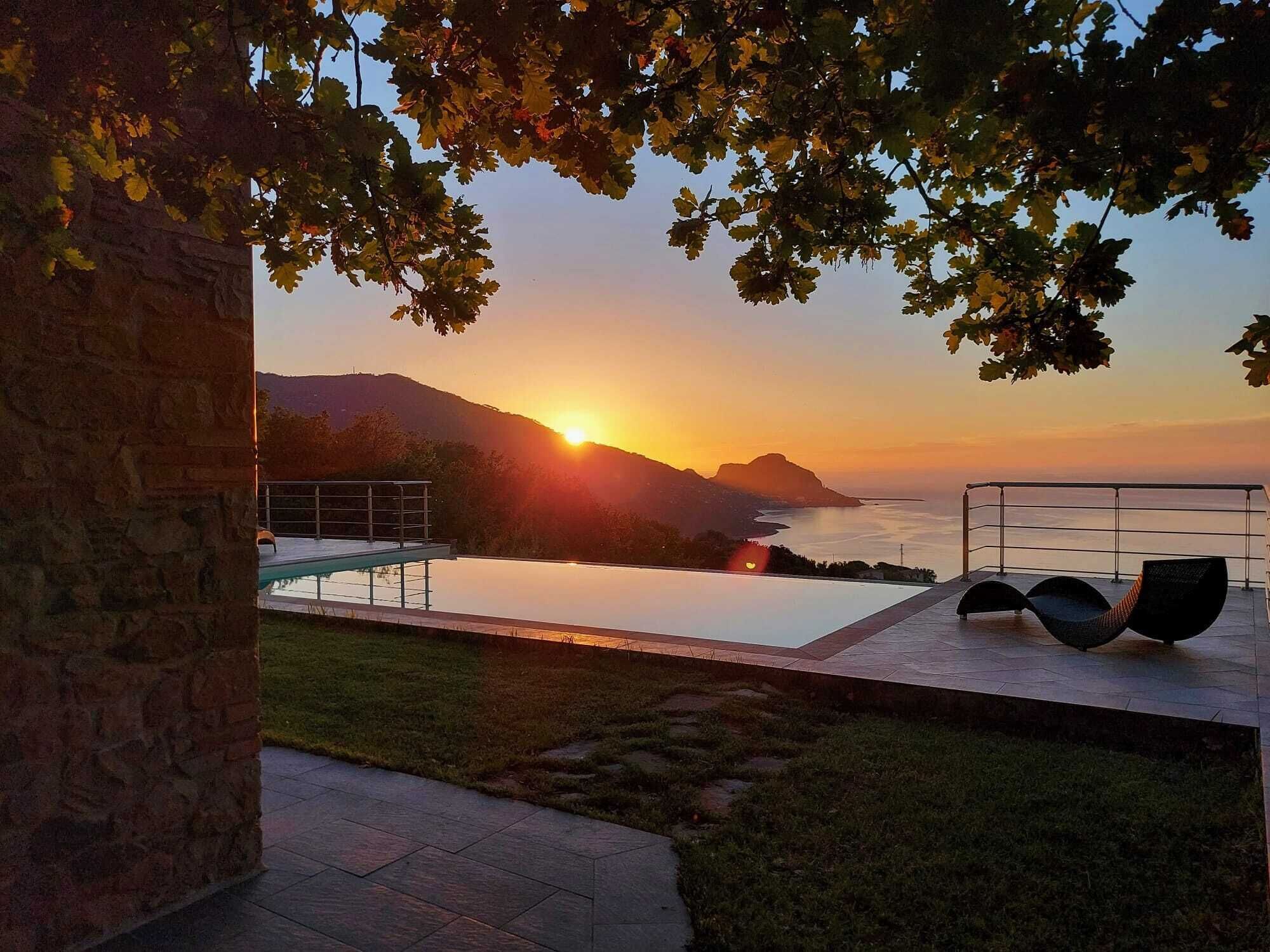 Zona junto a la piscina con vista al atardecer sobre el mar, un muro de piedra y una silla de descanso.
