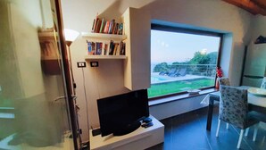 Una sala de estar moderna con un televisor de pantalla plana, estanterías y un gran ventanal con vistas a una piscina y follaje.