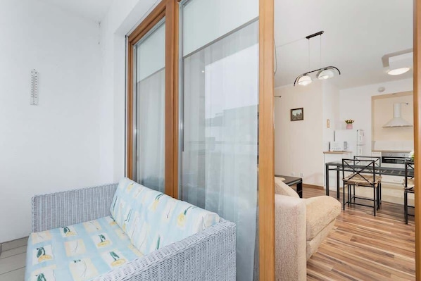 Ein modernes Wohnzimmer mit einem Rattan-Sofa, einem Holzespeisetisch und einem durch eine Glastür einsehbarer Küchenbereich.