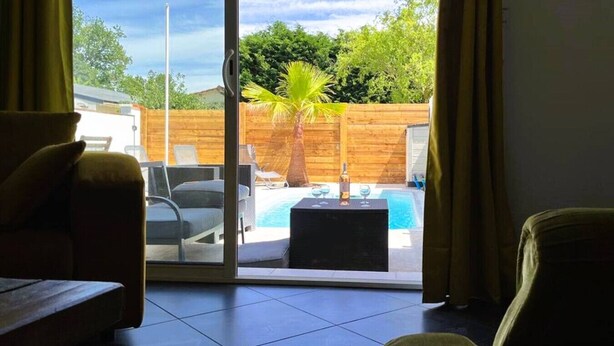 A room with a sliding glass door leading to a backyard with a pool, a palm tree, and a wooden fence. - Loft Nature Bien-être Piscine Chauffée Privée Tarn (Peyrole)