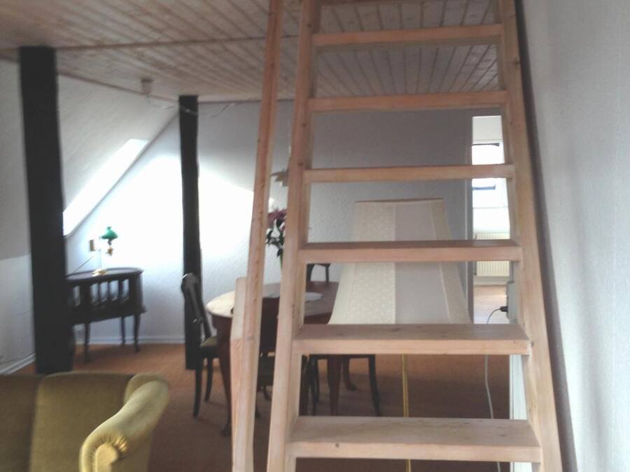 A wooden ladder in a room with a high ceiling, a dining table, and a green armchair.