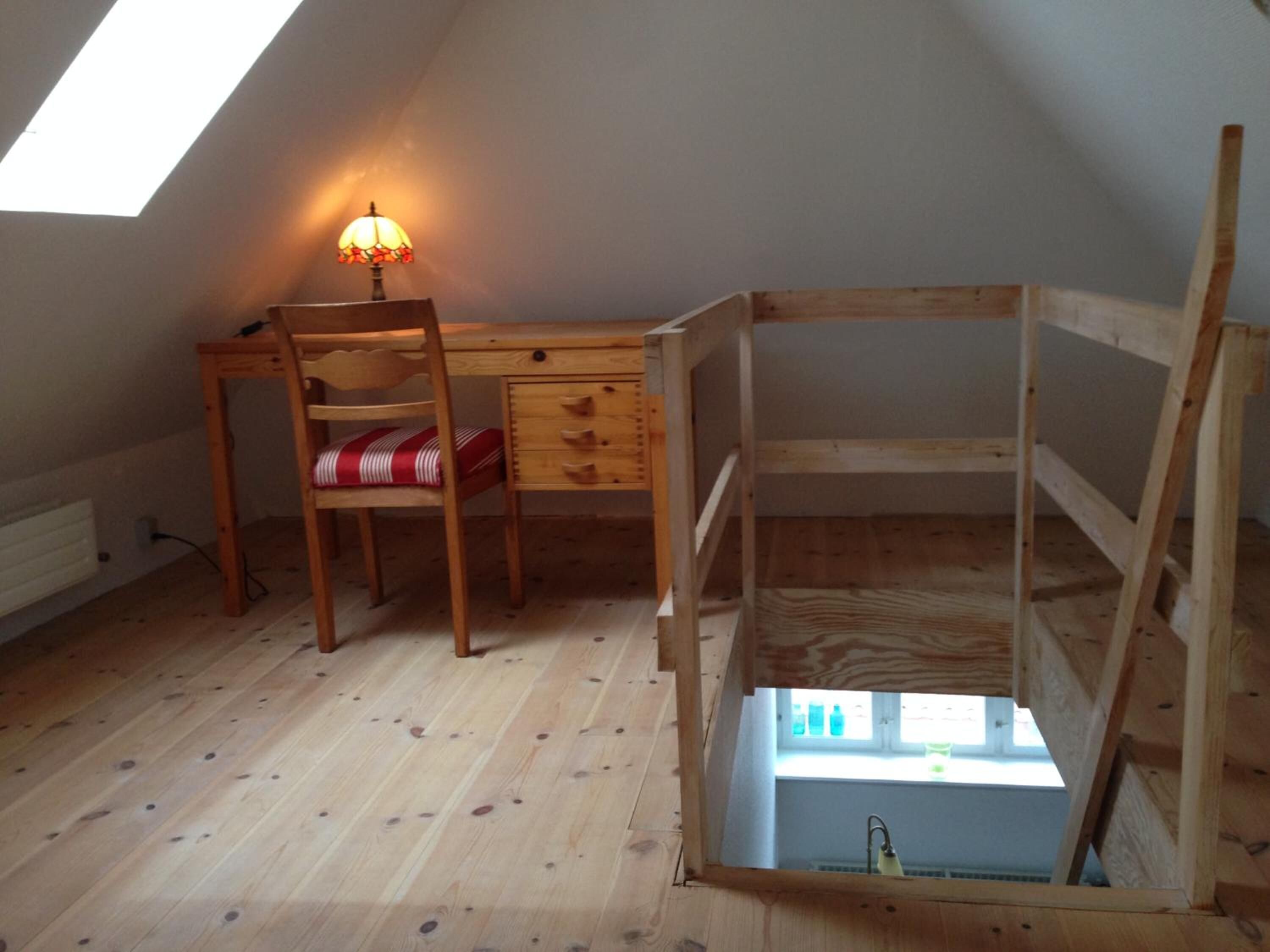 A small attic room with a slanted ceiling, a wooden desk with a chair, a lamp, and a wooden bunk bed.
