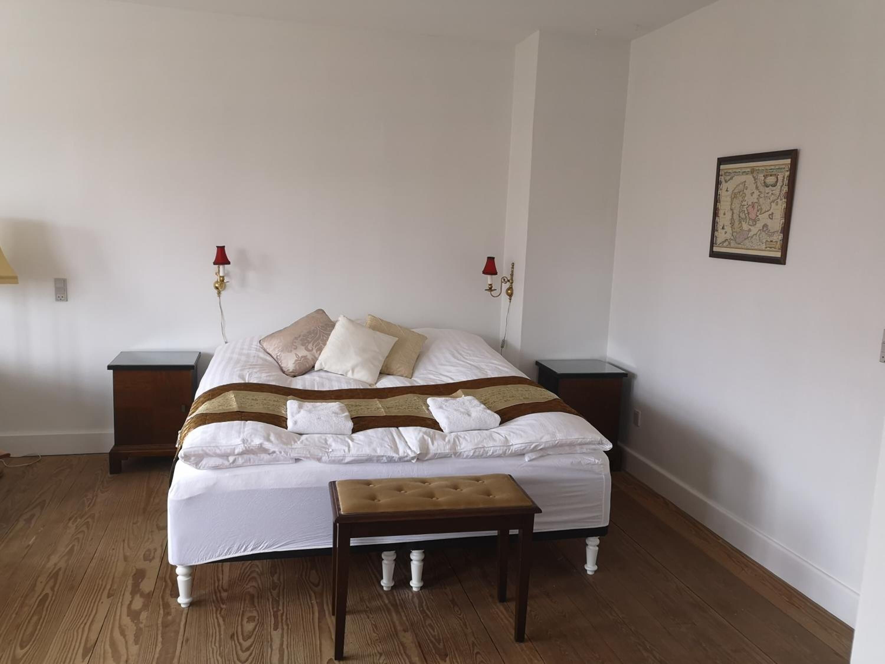 A neatly made bed with white linens, a brown and white striped blanket, and a matching bench. There are two bedside tables with lamps, and a framed picture on the wall.