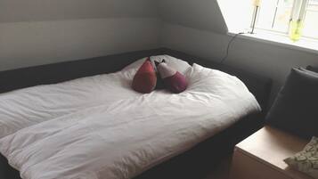 A single bed with white bedding and two decorative pillows, a bedside table with a lamp, and a window with a view of greenery.