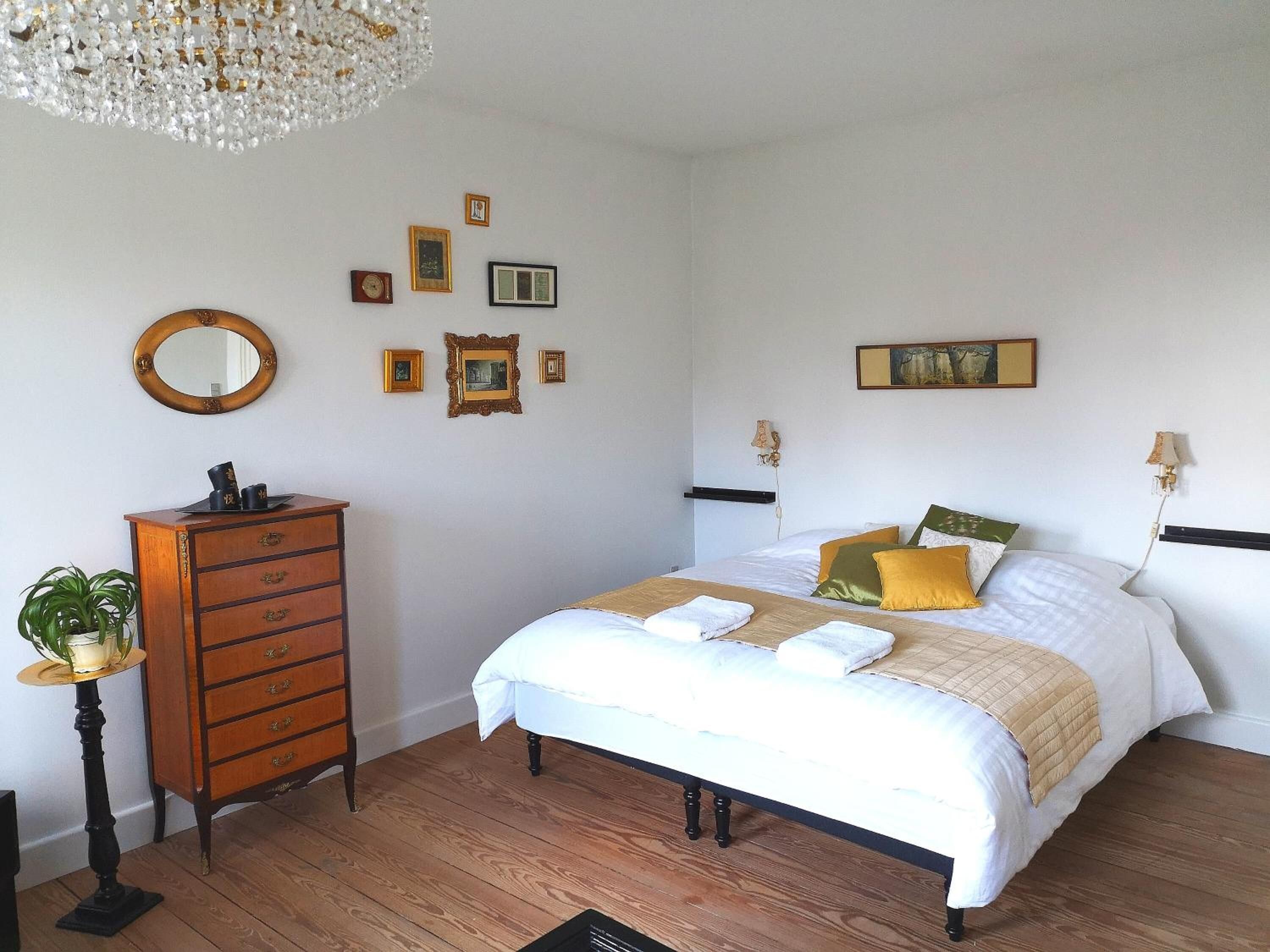 A bedroom with a wooden dresser, a bed with white bedding and multiple pillows, a chandelier, and several framed pictures on the wall.