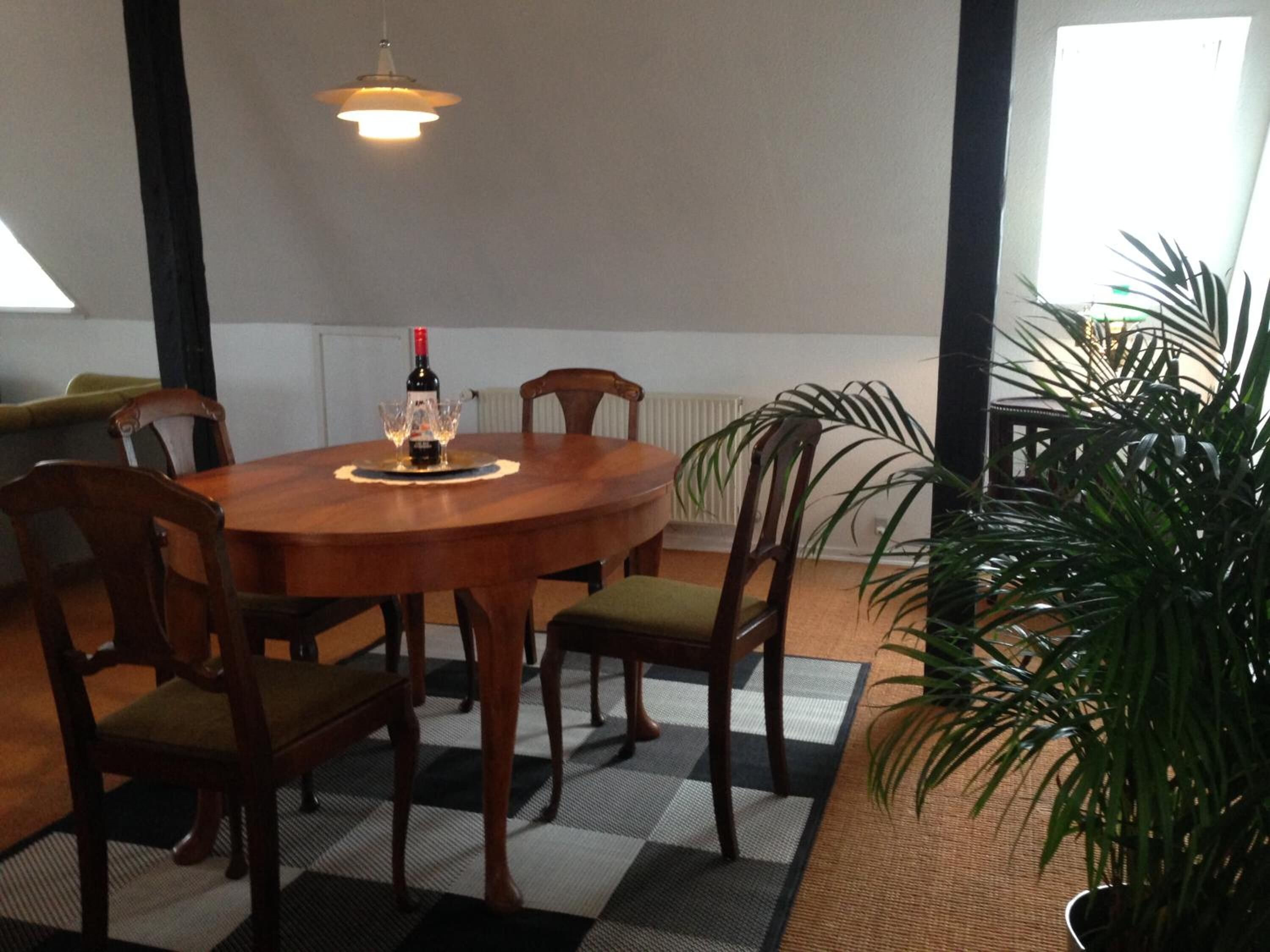 A dining area with a round wooden table, six chairs, a bottle of wine, and a lit pendant light.