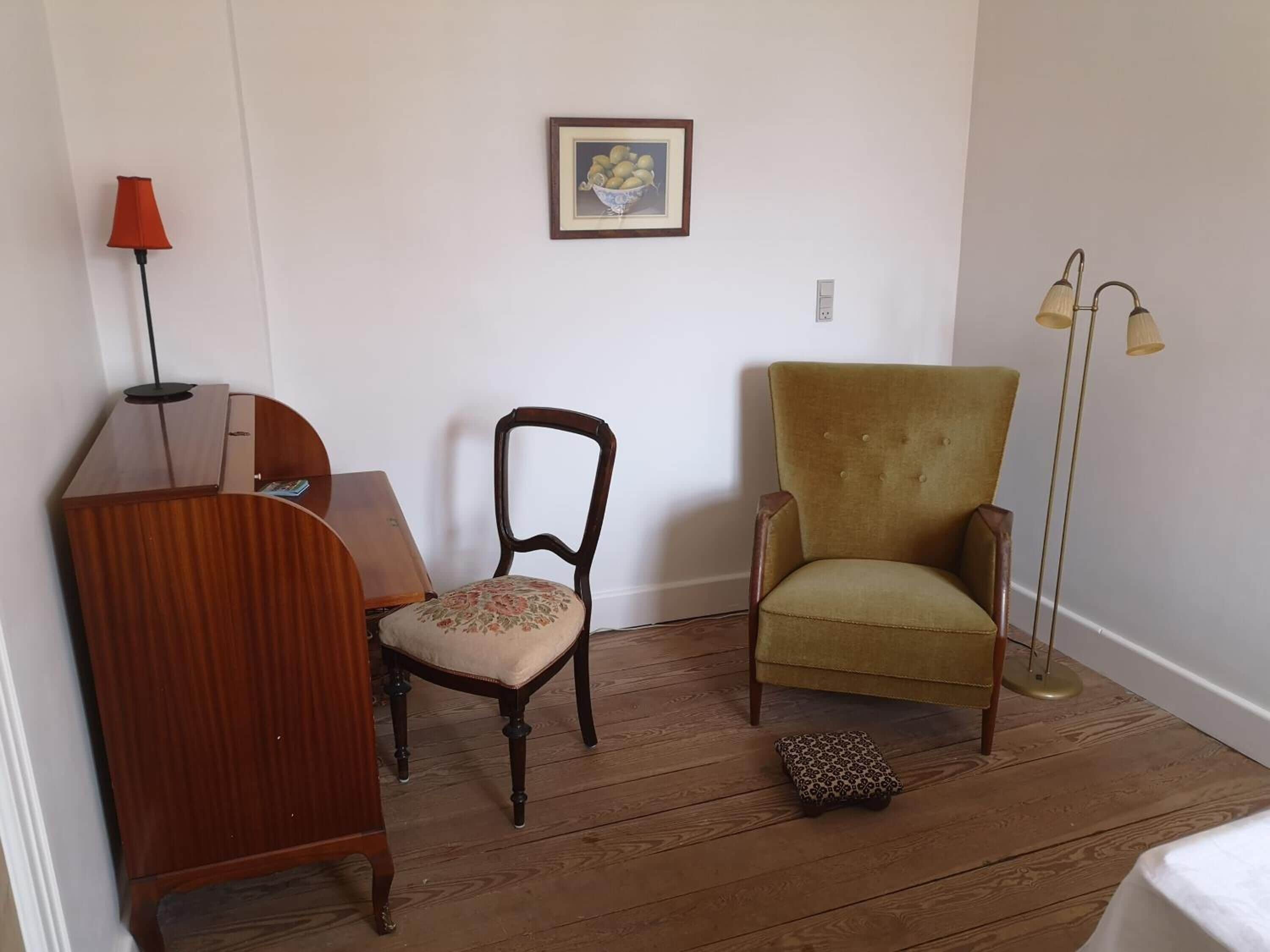 Una habitación con un escritorio de madera, una silla, un sillón verde, una lámpara de pie y un cuadro en la pared.