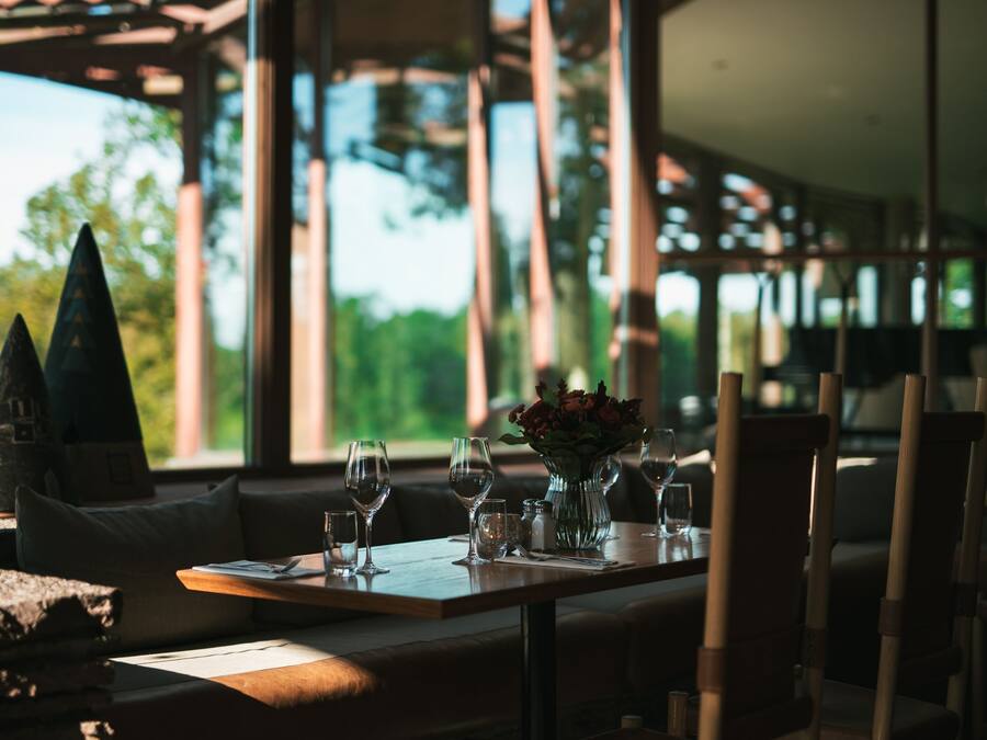 A dining area with a table set for two, a vase with flowers, and a view of trees through large windows.