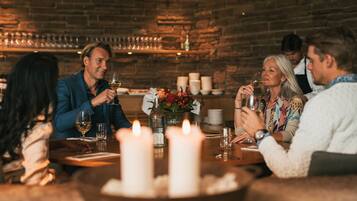 Un grupo de personas cenando en una mesa con velas, copas de vino y un muro de ladrillo al fondo.