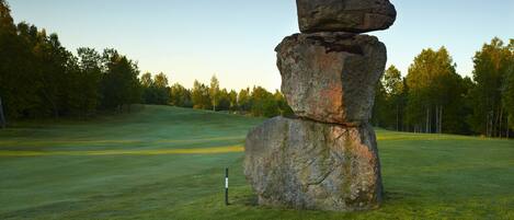 A large, multi-tiered rock formation in a grassy field with trees in the background.