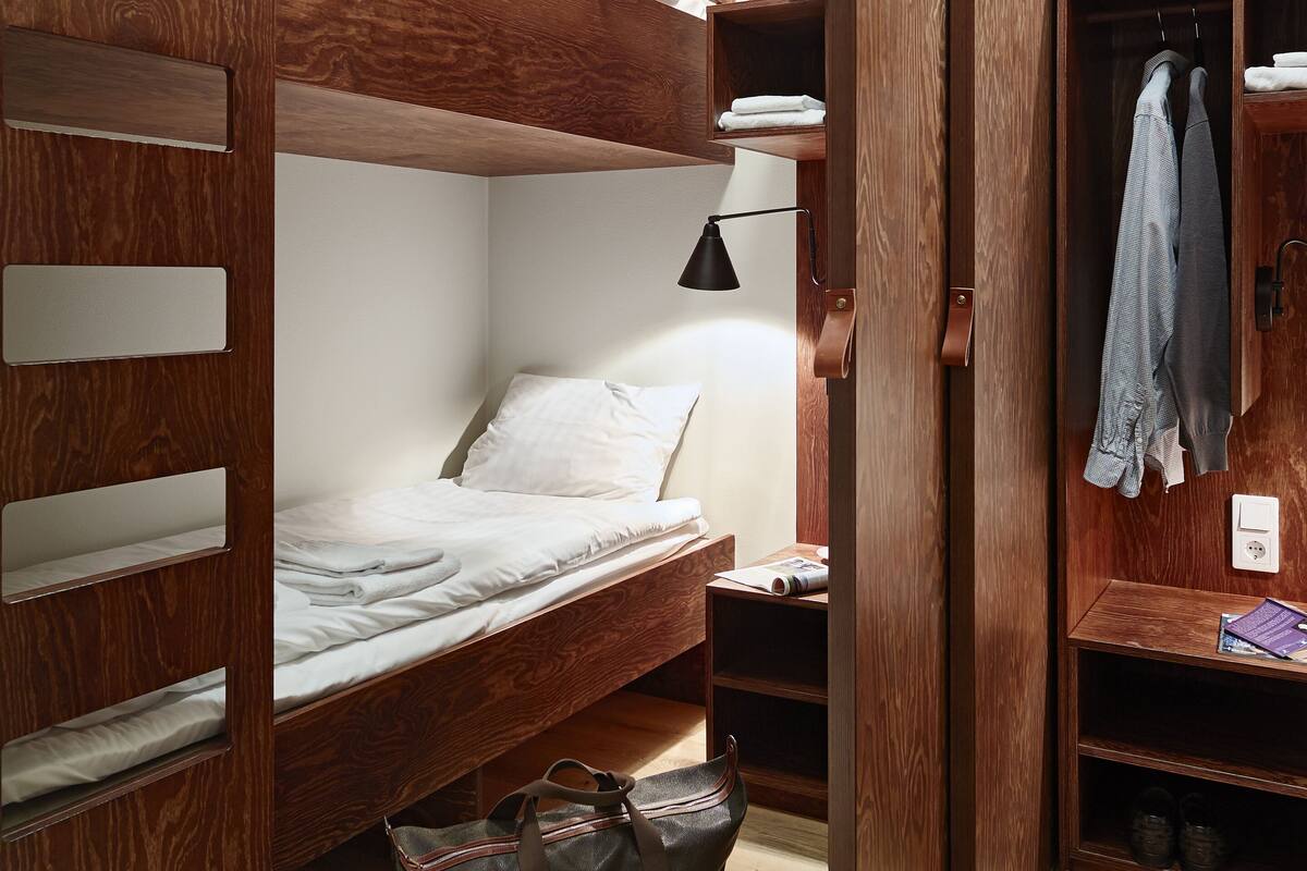 Una cama nido con cabecero de madera y estantes, una mesita de noche con lámpara, y una bolsa en el suelo.
