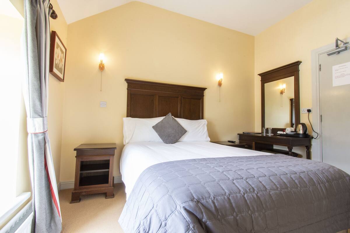 A hotel room with a large bed, a bedside table, a nightstand, a dresser, a mirror, and a door with a sign.