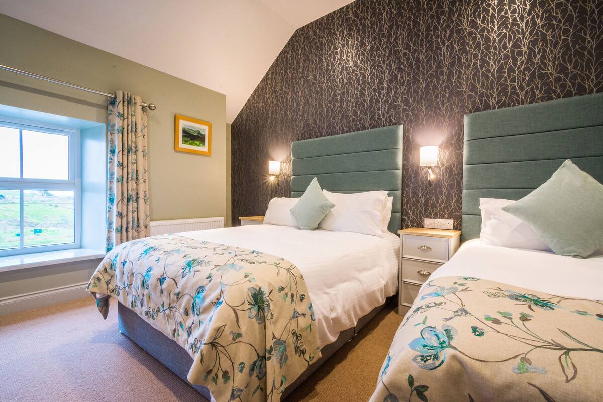 A hotel room with two beds, a window with curtains, a wall with a tree pattern, and a nightstand with a lamp.
