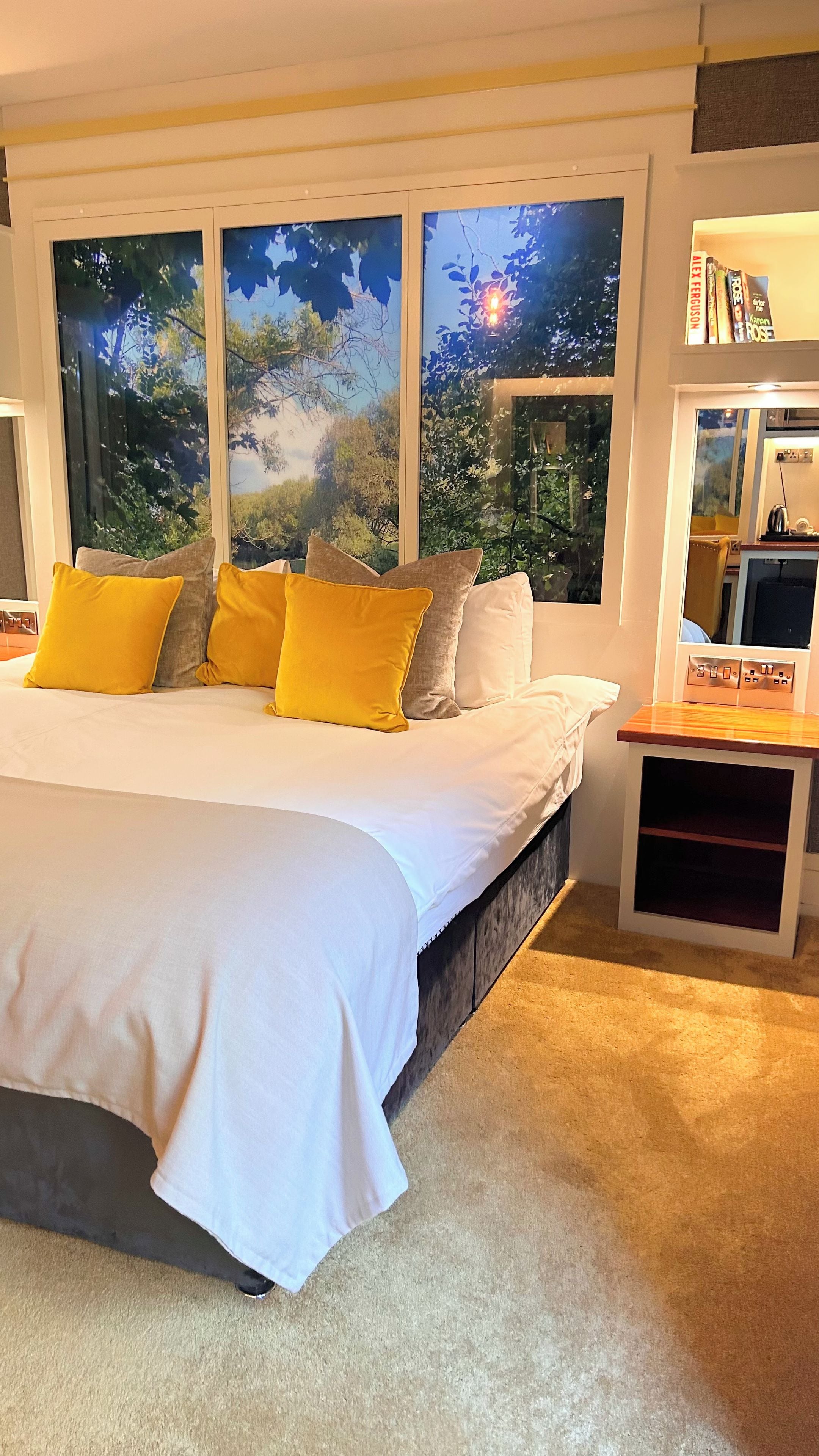 A bedroom with a large bed, multiple pillows, a nightstand, a bookshelf, and a window with a view of trees.