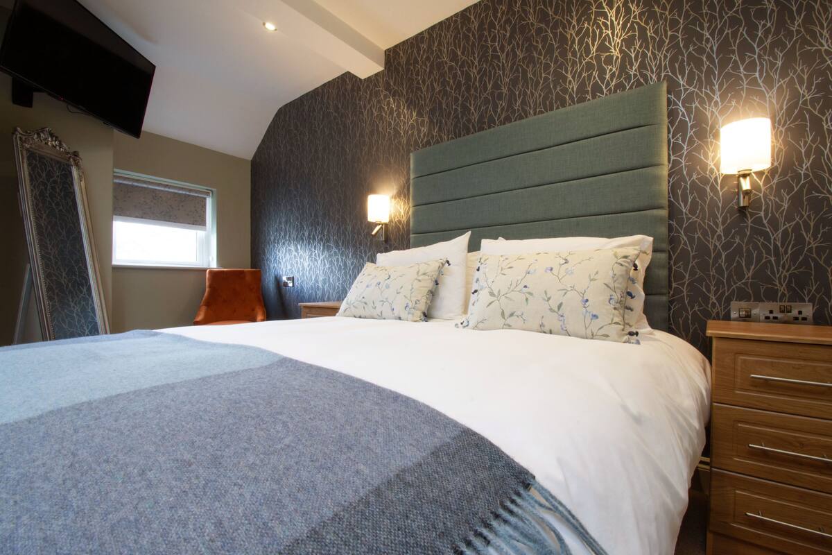 A bedroom with a large bed, two bedside lamps, a wooden dresser, and a television mounted on the wall.