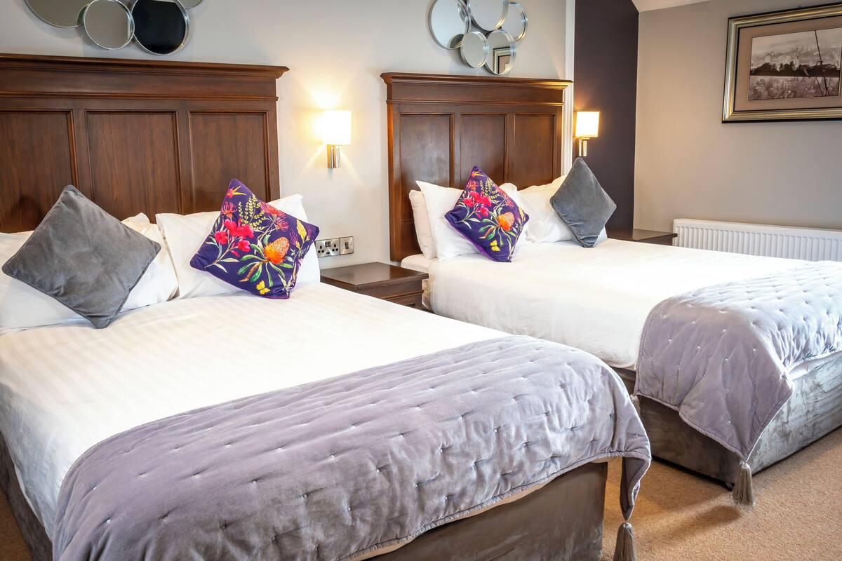 A hotel room with two beds, each with a headboard, a nightstand, a lamp, and a wall-mounted mirror.