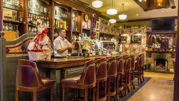 A bar area with a bartender, bar stools, shelves with bottles, and a fireplace. - Nevins Inn (Newport)