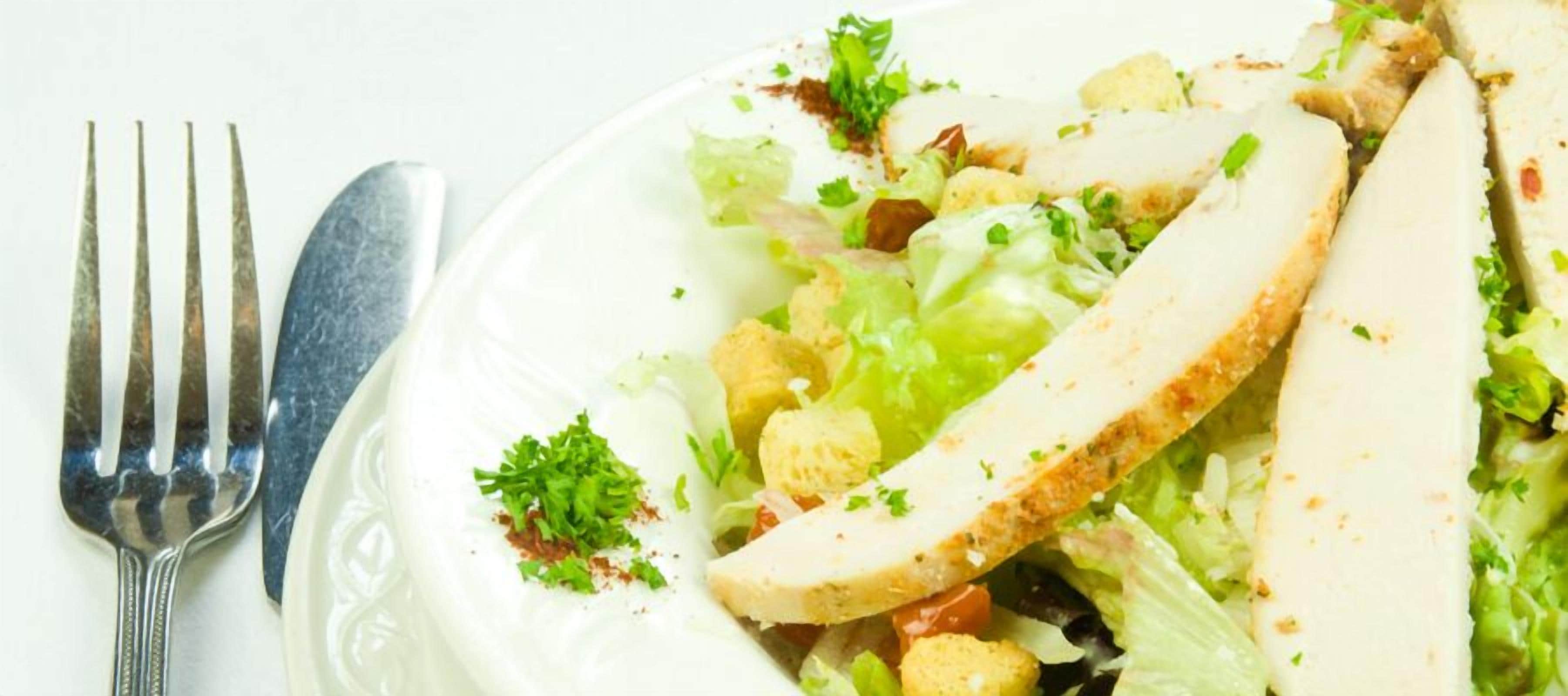 Une assiette de nourriture comprenant de la laitue, des croûtons et une tranche de viande blanche, accompagnée d’une fourchette.
