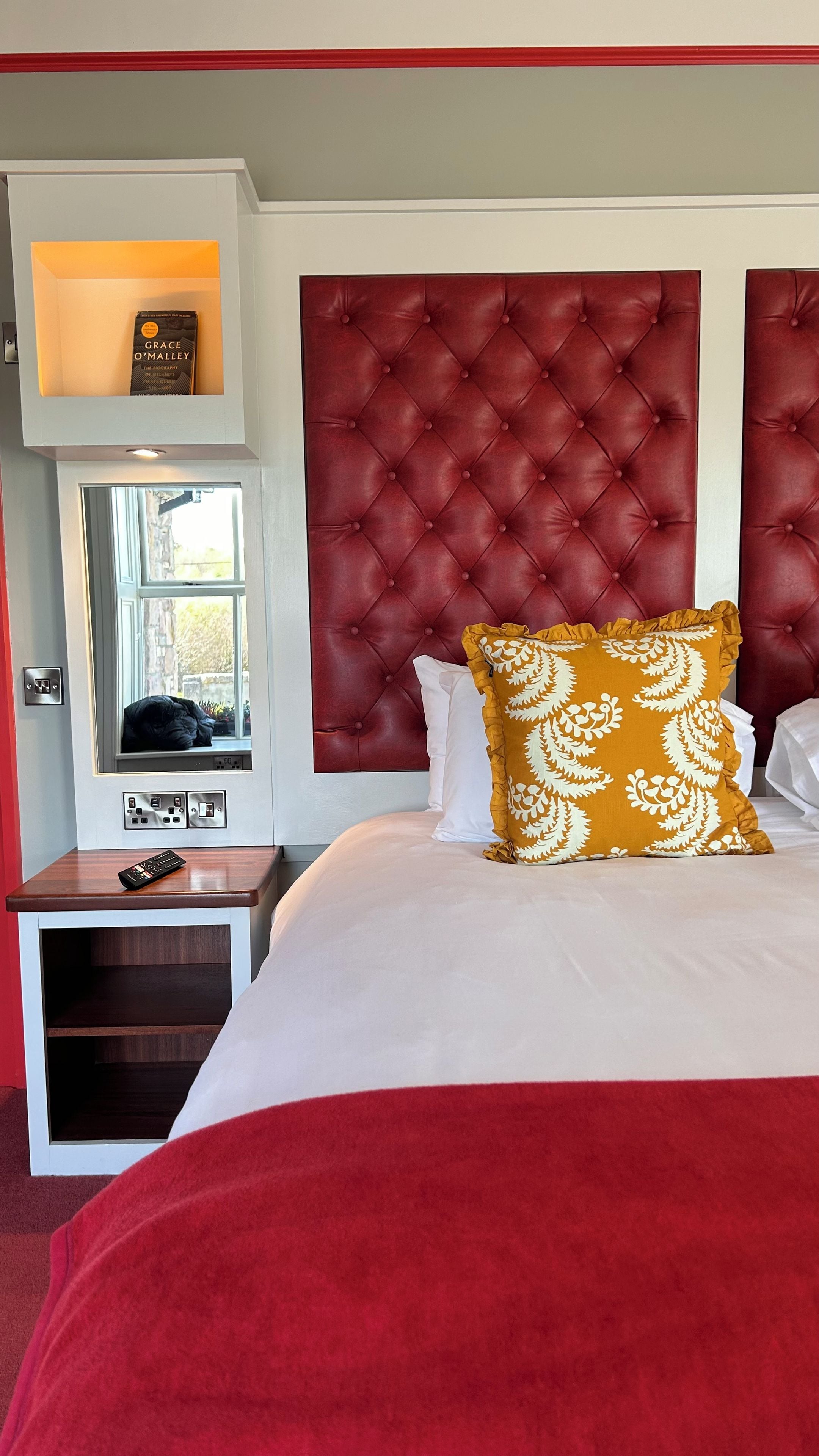 A hotel room with a tufted headboard, a window with a view, a bedside table with a remote control, and a bed with white and red bedding.