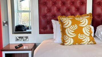 A hotel room with a tufted headboard, a window with a view, a bedside table with a remote control, and a bed with white and red bedding.