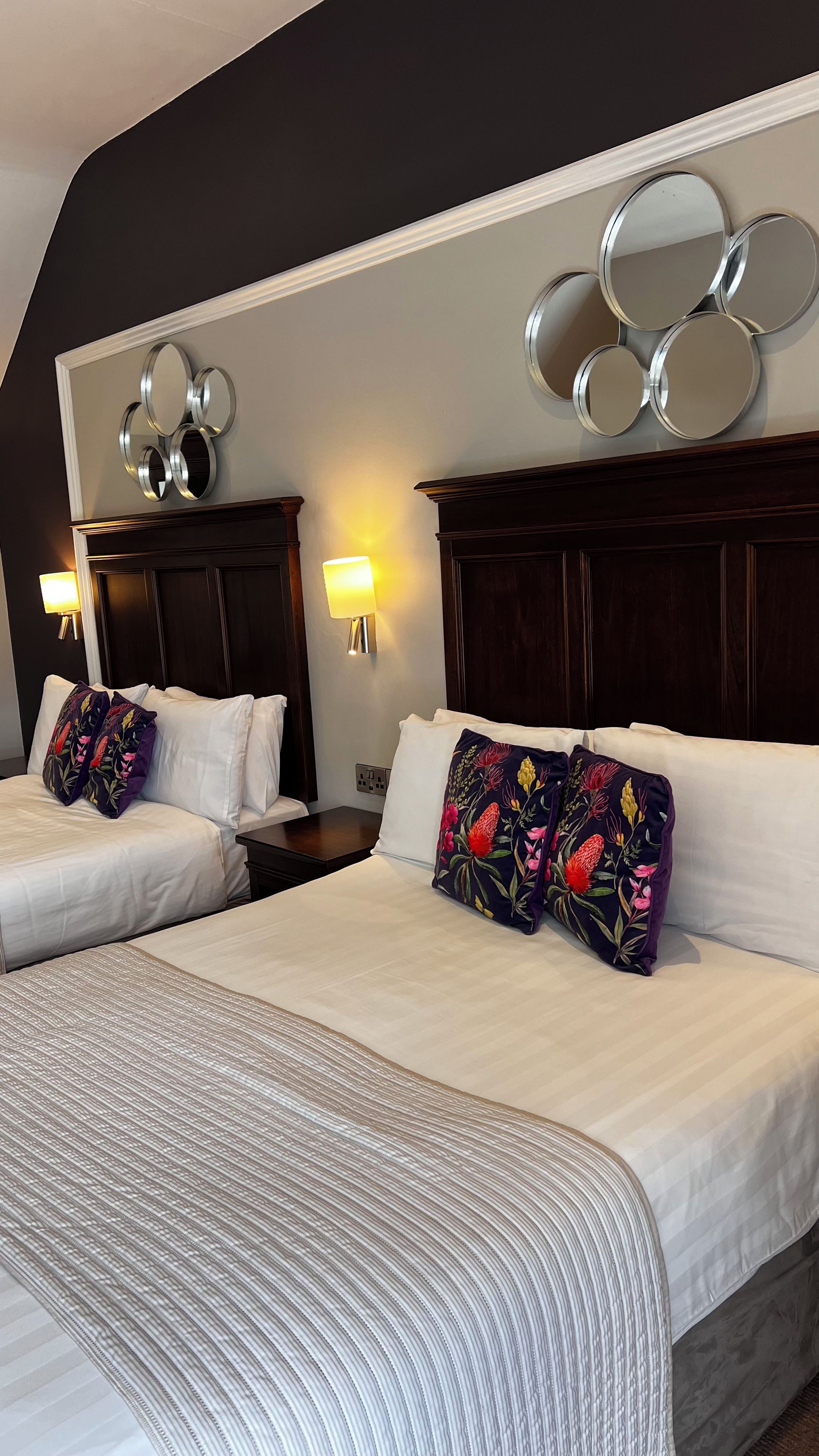 A hotel room with two beds, each with white linens and floral-patterned pillows. There are wall-mounted mirrors above the beds and a wall sconce with a warm light.
