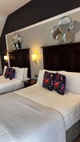 Habitación de hotel con dos camas, cada una con ropa blanca y almohadas estampadas con motivos florales. Hay espejos en la pared sobre las camas y un aplique con luz cálida.