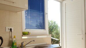 A kitchen with wooden cabinets, a window with blinds, a sink, and a door leading outside. - Studio apartment GIRO (Omis)
