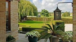 A brick building with a balcony overlooking a well-manicured garden with a weeping willow, a lantern, and potted plants. - B&B Bertem Natuur (Bertem)