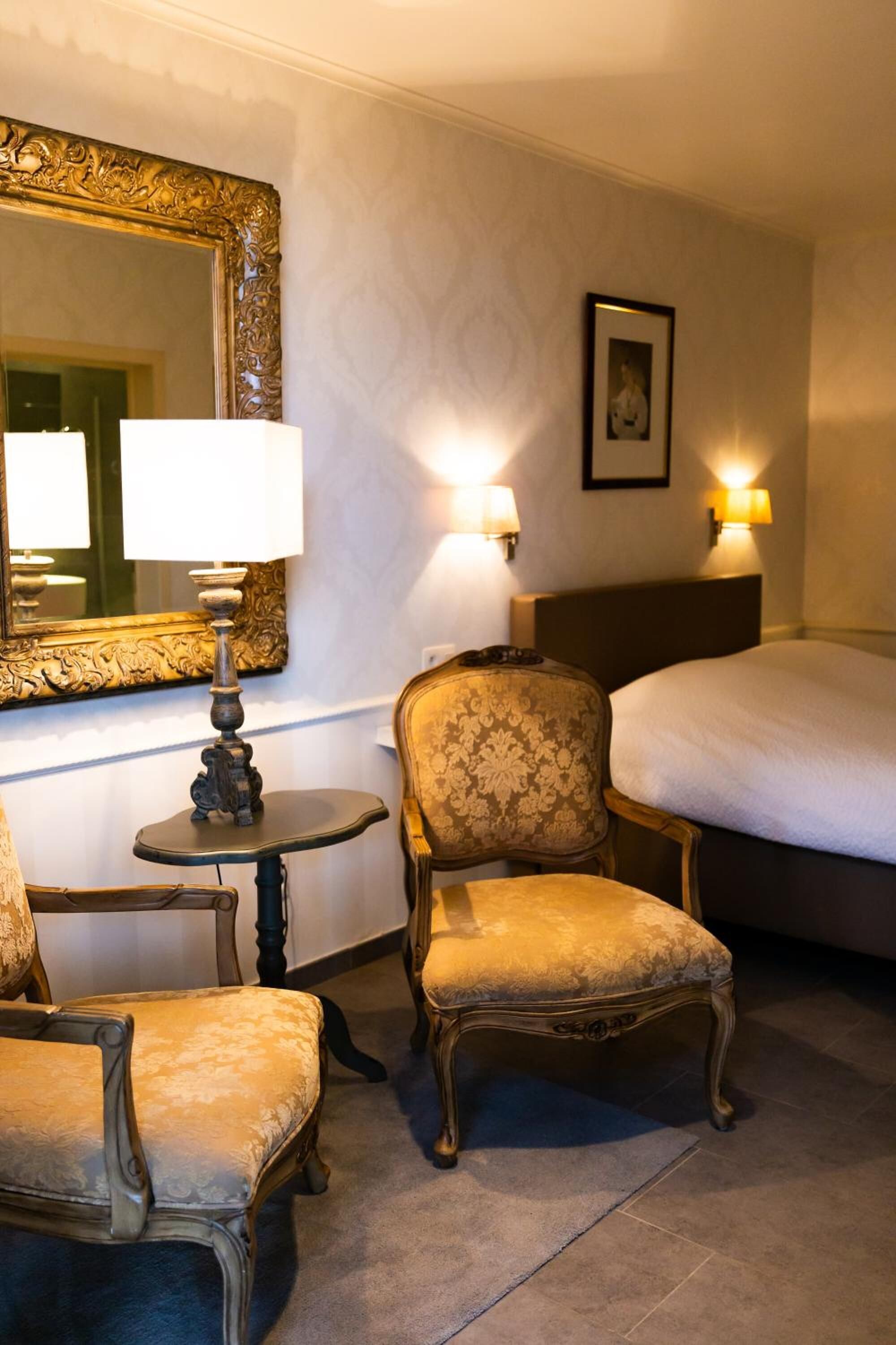 A hotel room with a bed, two chairs, a small table with a lamp, a mirror, and wall-mounted lamps.