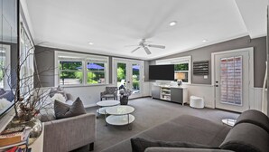 A spacious living room with a ceiling fan, large windows, a flat-screen TV, and a kitchen area with a countertop and cabinets. - Huge Backyard, Large Living Room with Dining Area, Large Den with Built-In... (Southampton)